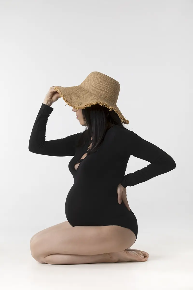 Futura mamma di profilo seduta con body nero e cappello di paglia, ritratto premaman in studio a Milano