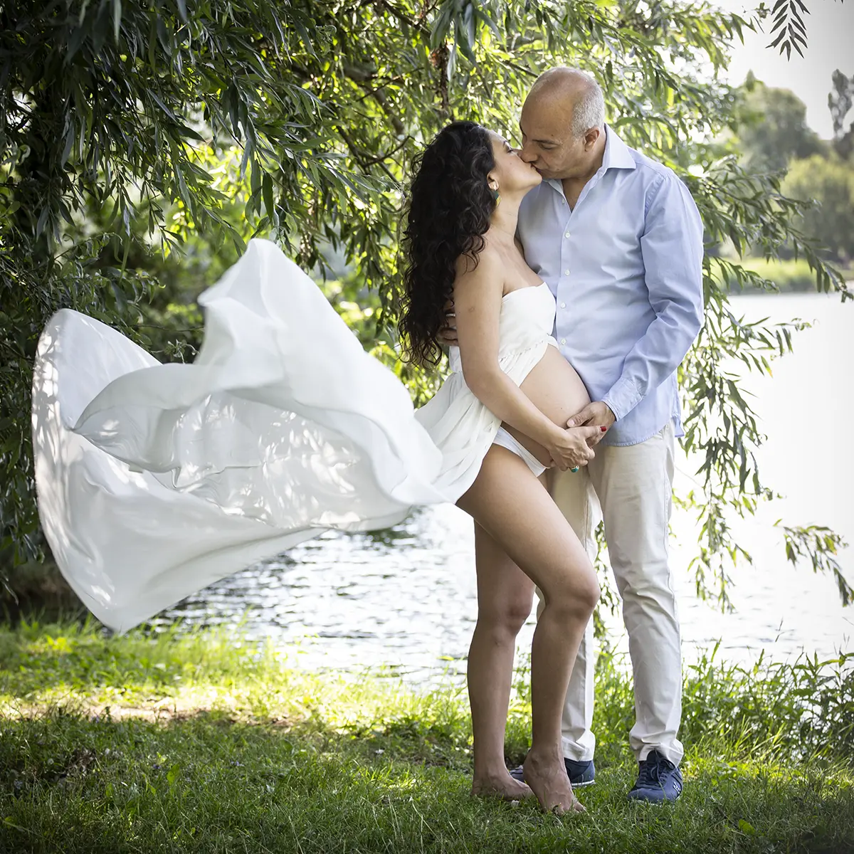 Coppia in attesa che si bacia durante un book fotografico gravidanza all'aperto a Milano, abito bianco vicino al lago